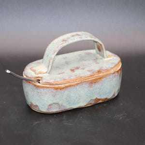 May include: An oval ceramic butter dish with a matching lid featuring a curved handle. The piece has a light blue, mottled glaze with visible brown clay edges and a small metal spoon resting in a side notch on the lid.