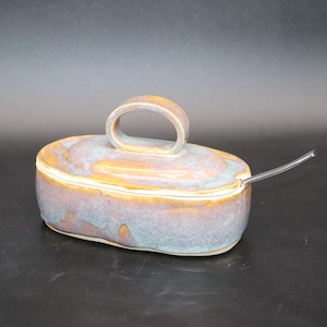 May include: An oval ceramic butter dish with a lid featuring a prominent arched handle. The glaze is a blend of muted blue, lavender, and warm golden-brown tones. A small, clear, transparent plastic spoon is inserted into the side of the dish.