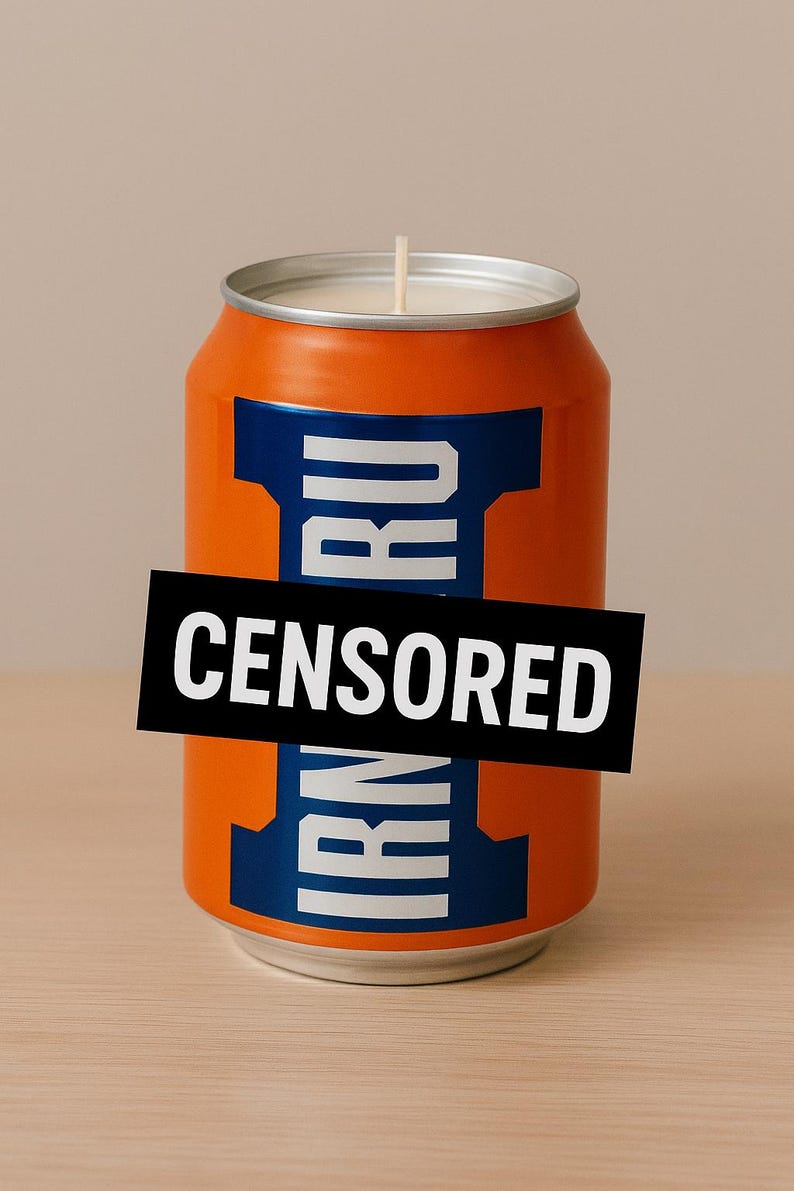 May include: Orange candle in the shape of a soda can, with blue and white accents. The text "IRN RU" is partially visible, with a black rectangle obscuring the rest of the text. A white wick is centered on the top.