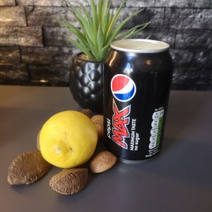 May include: A black can of Pepsi Max with the text "Maximum Taste No Sugar" on the side. The can is sitting on a gray surface with a lemon and several brown nuts.