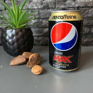 May include: A black can of Pepsi Max with the words "no caffeine" and "maximum taste no sugar" printed on it. The can is sitting on a gray surface with a few brown nuts.