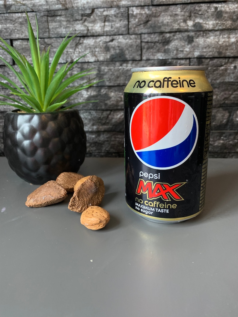 May include: A black can of Pepsi Max with the words "no caffeine" and "maximum taste no sugar" printed on it. The can is sitting on a gray surface with a few brown nuts.