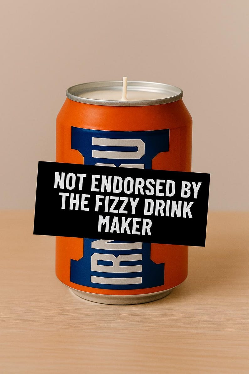 May include: Orange and blue candle in the shape of a soda can with a white wick. A black rectangle with white text that reads "NOT ENDORSED BY THE FIZZY DRINK MAKER" is adhered to the front. The candle sits on a wooden surface.