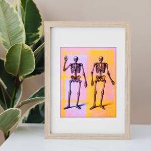 Puede incluir: Obra de arte enmarcada con dos ilustraciones de esqueletos. Un esqueleto está coloreado en tonos morados, el otro en tonos dorados. El fondo es una mezcla de cuadrados rosas, amarillos y azules. La obra está en un marco de madera clara.
