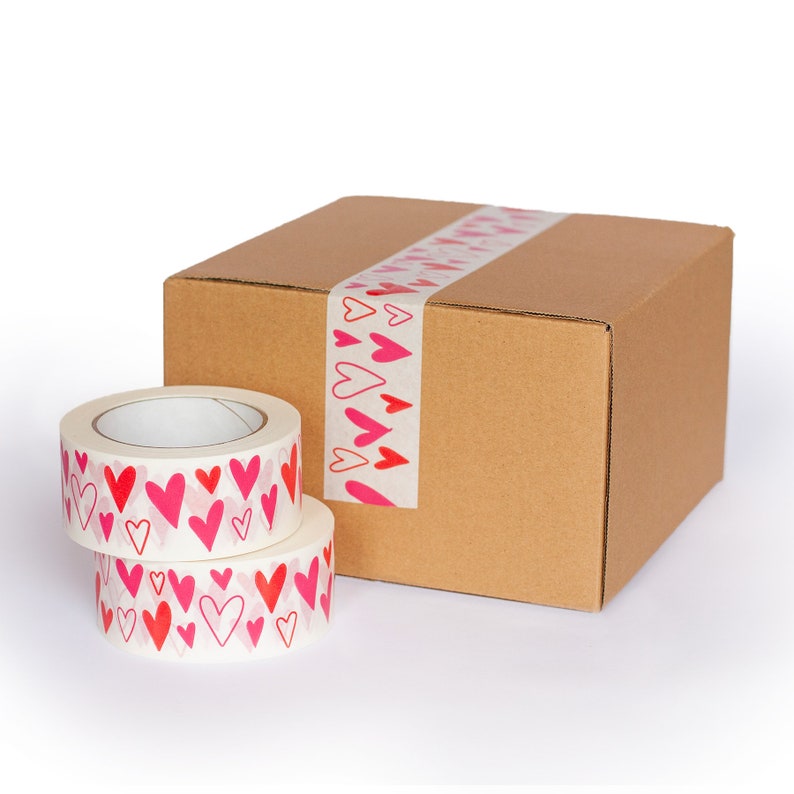Puede incluir: Dos rollos de cinta adhesiva decorativa con patrones de corazones rojos y rosas, apilados frente a una caja de cart&oacute;n sellada con cinta a juego. La cinta es blanca con un dise&ntilde;o de coraz&oacute;n repetido, ideal para envolver regalos o manualidades.
