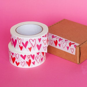 Puede incluir: Dos rollos de cinta adhesiva decorativa blanca con un patr&oacute;n repetido de corazones rojos y rosas. Una caja de cart&oacute;n marr&oacute;n est&aacute; pegada con la misma cinta con estampado de corazones. El fondo es rosa liso.