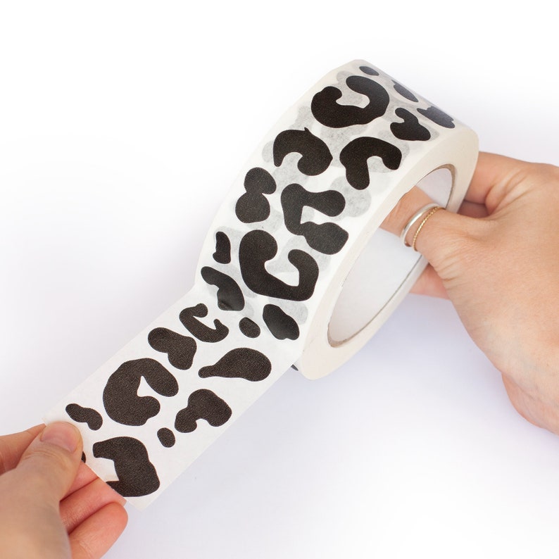 May include: A roll of white washi tape with a black leopard print design. The tape is being unrolled, revealing the pattern.