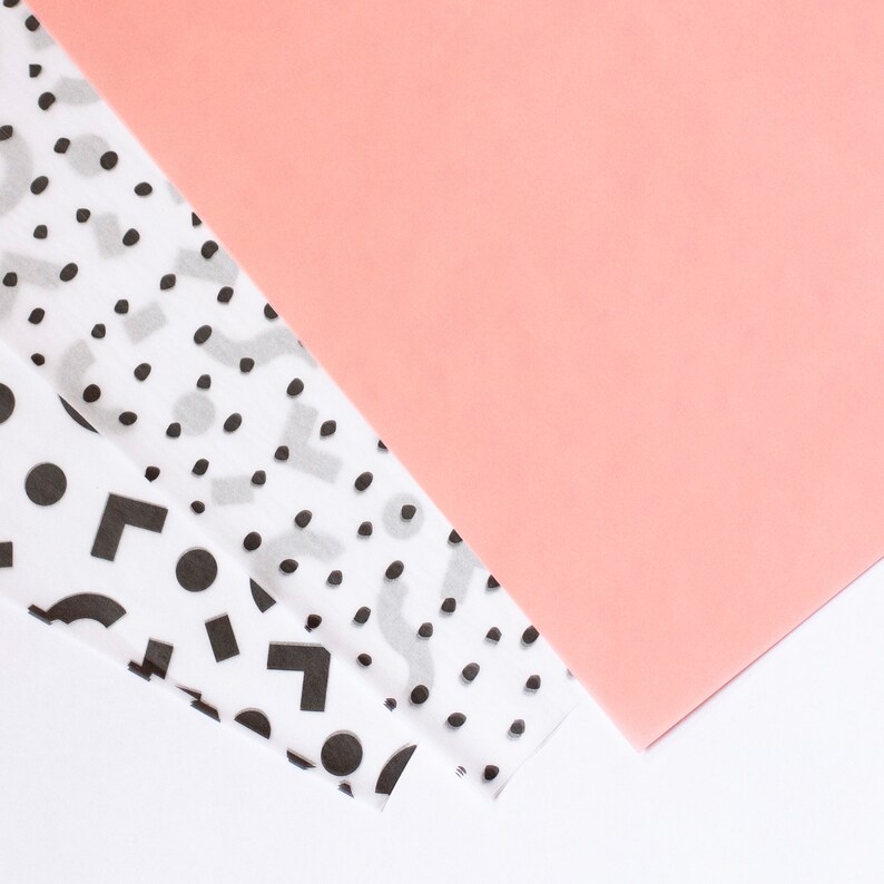 May include: Two sheets of white tissue paper with black geometric patterns and polka dots, layered on top of a pink sheet of paper.