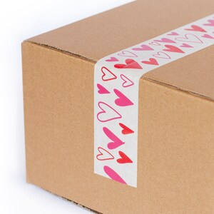 Puede incluir: Una caja de cart&oacute;n marr&oacute;n sellada con cinta adhesiva blanca con un patr&oacute;n repetido de corazones rojos y rosas. Los corazones var&iacute;an en tama&ntilde;o y estilo, algunos delineados y otros rellenos.