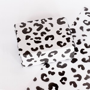 May include: A white gift box with a black leopard print design. The box is wrapped in white tissue paper with the same leopard print design.