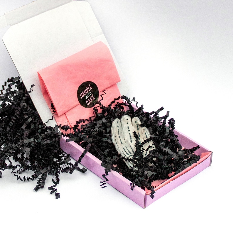 May include: A small, light purple box with black shredded paper holds a white and gray decorative object. A pink tissue paper package with a "HANDLE WITH CARE" sticker sits inside the box. The box is inside a larger white box.