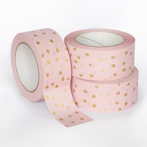 K&ouml;nnte beinhalten: Drei Rollen rosa Washi-Tape mit goldenen Punkten. Das Band ist aufgerollt und die Enden sind abgewickelt, so dass das Muster sichtbar ist.