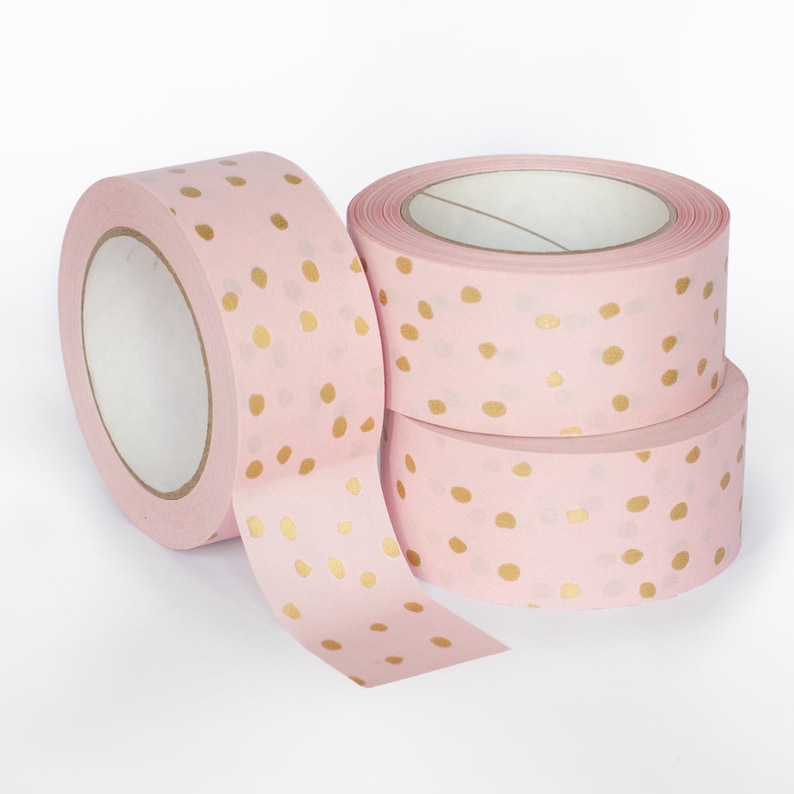K&ouml;nnte beinhalten: Drei Rollen rosa Washi-Tape mit goldenen Punkten. Das Band ist aufgerollt und die Enden sind abgewickelt, so dass das Muster sichtbar ist.