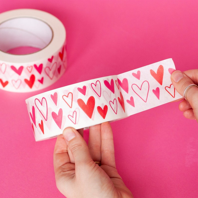 Puede incluir: Un rollo de cinta adhesiva decorativa con un patr&oacute;n repetido de corazones rojos y rosas. La cinta se est&aacute; desenrollando, revelando el dise&ntilde;o de coraz&oacute;n. El fondo es de color rosa s&oacute;lido.