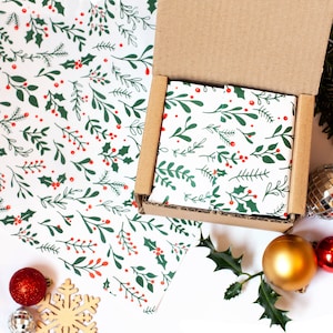 May include: A small cardboard box with a white and green holly pattern wrapping paper. The box is open and the inside is lined with the same wrapping paper. The box is surrounded by Christmas decorations, including red and gold ornaments and a snowflake.