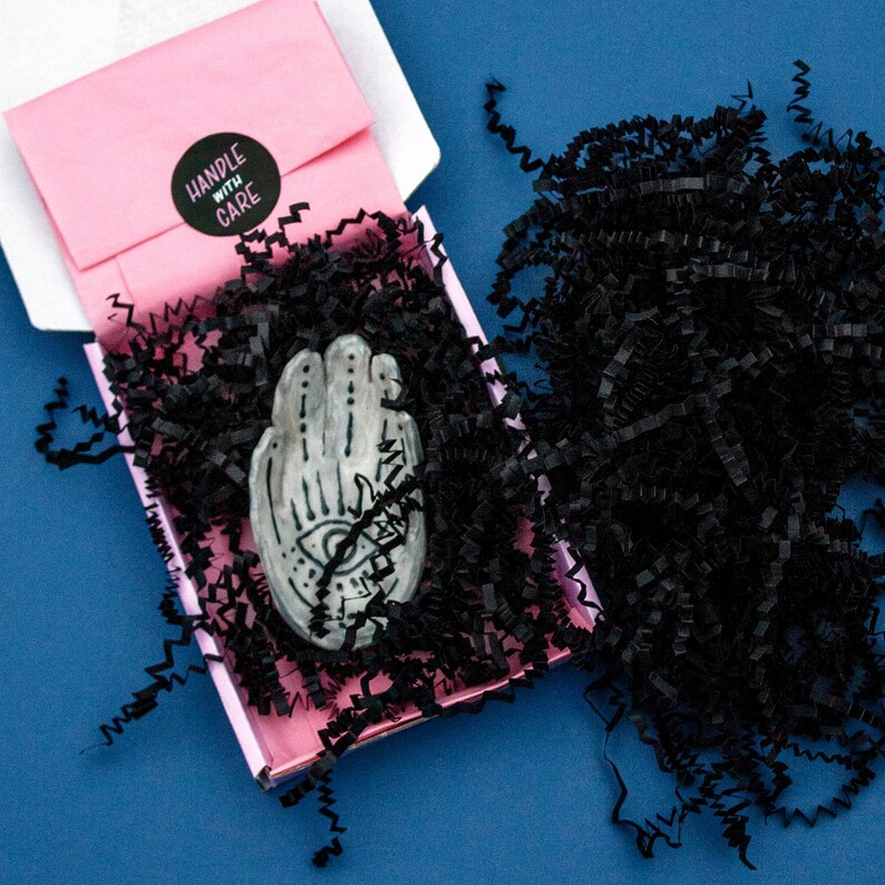 May include: A gray ceramic hand with an eye design, packaged in a pink box with black crinkle paper. The box includes a pink envelope with a "HANDLE WITH CARE" sticker.