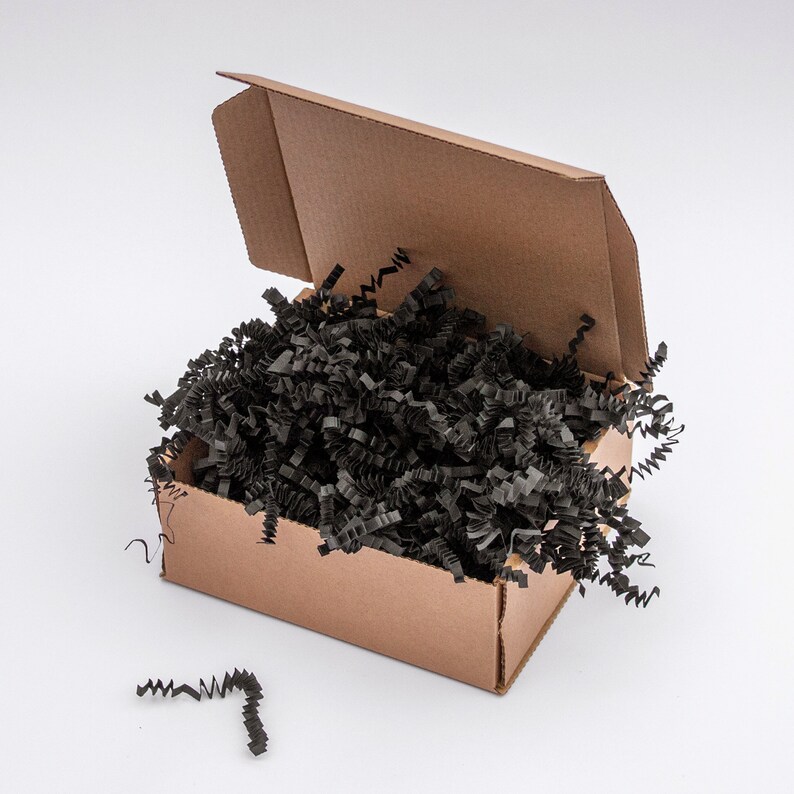 May include: A brown cardboard box with the lid partially open, filled with black crinkle cut paper. A few pieces of the black paper are scattered outside the box. The box is on a white background.