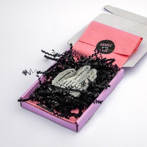May include: A small, open pink box containing a white and gray hand-shaped sculpture, nestled in black paper shreds. A pink envelope with a "HANDLE WITH CARE" sticker is inside the box. The box is on a white surface.
