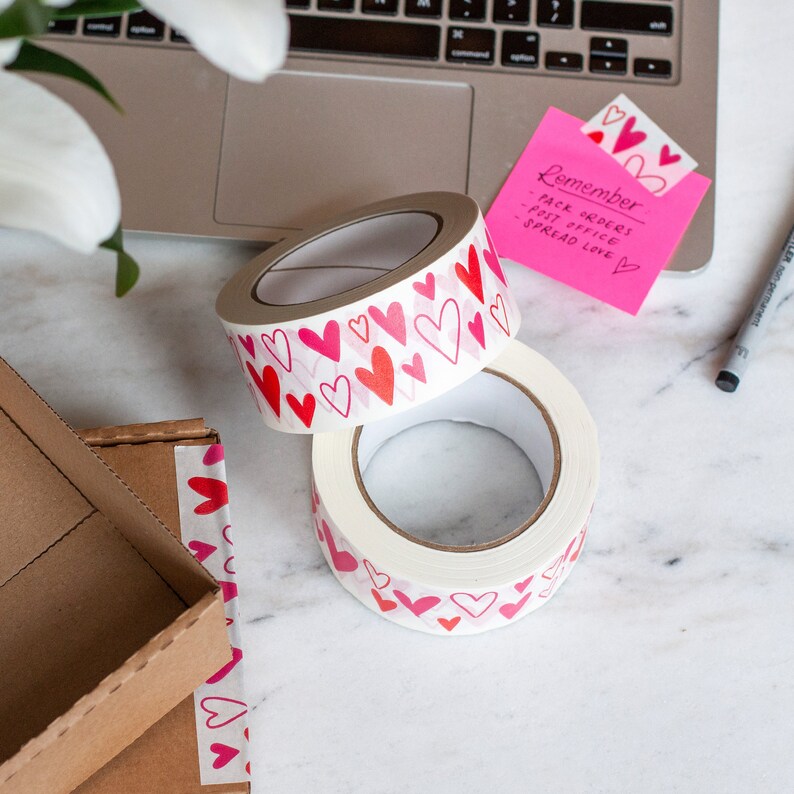 Puede incluir: Dos rollos de cinta adhesiva decorativa con corazones rojos y rosas sobre fondo blanco. Una nota adhesiva rosa con la palabra "Remember" y otro texto es visible. Un port&aacute;til y un bol&iacute;grafo est&aacute;n en el fondo. Para envolver regalos.