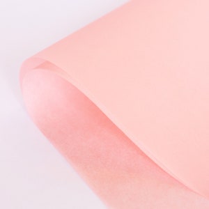 May include: A close-up of a sheet of light pink tissue paper. The paper is folded in half, and the edges are slightly curled.