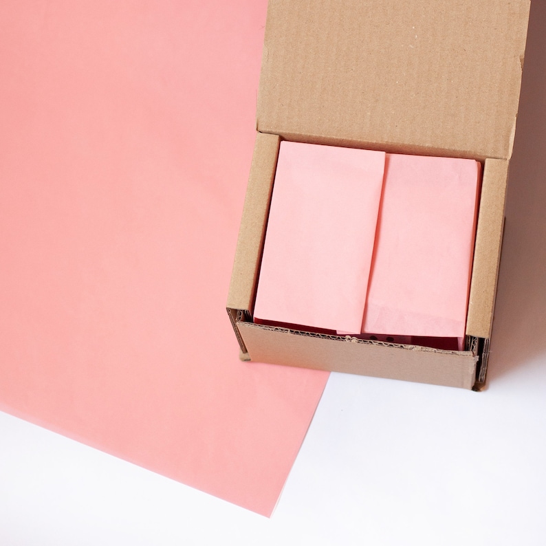 May include: A cardboard box with the top open, revealing pink tissue paper inside. The box is sitting on a pink surface.