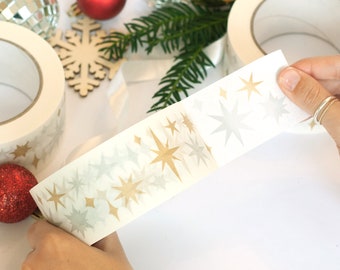 Puede incluir: Cinta adhesiva decorativa con un patrón de estrellas doradas y plateadas. La cinta se está desenrollando, revelando el diseño. El fondo incluye adornos navideños y vegetación, sugiriendo un tema festivo.