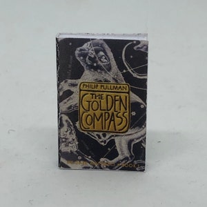 Puede incluir: Una cubierta de libro negra y dorada con un dise&ntilde;o de constelaci&oacute;n. El t&iacute;tulo "The Golden Compass" est&aacute; en letras doradas. El autor, Philip Pullman, est&aacute; impreso en letras doradas m&aacute;s peque&ntilde;as debajo del t&iacute;tulo.