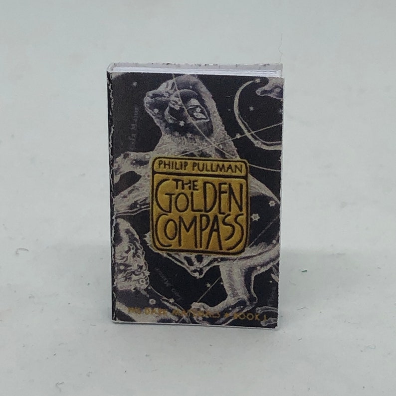 Puede incluir: Una cubierta de libro negra y dorada con un dise&ntilde;o de constelaci&oacute;n. El t&iacute;tulo "The Golden Compass" est&aacute; en letras doradas. El autor, Philip Pullman, est&aacute; impreso en letras doradas m&aacute;s peque&ntilde;as debajo del t&iacute;tulo.