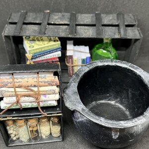Puede incluir: Una escena de boticario de brujas en miniatura con un caldero negro, un cofre de madera con libros y un pequeño armario con botellas de pociones. Los libros incluyen títulos como "Green Witchcraft".