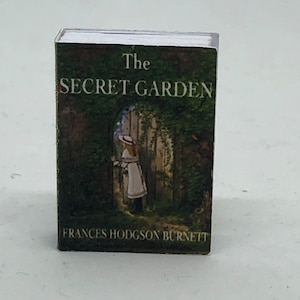 Puede incluir: Un libro en miniatura titulado "El jard&iacute;n secreto" de Frances Hodgson Burnett. La portada presenta una escena de jard&iacute;n verde con una ni&ntilde;a con un vestido blanco de pie junto a una puerta.