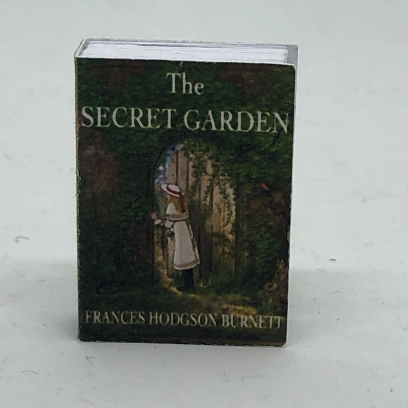 Puede incluir: Un libro en miniatura titulado "El jard&iacute;n secreto" de Frances Hodgson Burnett. La portada presenta una escena de jard&iacute;n verde con una ni&ntilde;a con un vestido blanco de pie junto a una puerta.