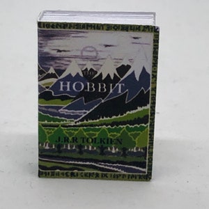 Puede incluir: Un libro de bolsillo vintage con una portada verde, azul y blanca que presenta una escena de paisaje con monta&ntilde;as y el t&iacute;tulo "El Hobbit" de J.R.R. Tolkien.