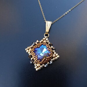 May include: A gold chain necklace with a square pendant featuring a blue crystal surrounded by gold and black beads.