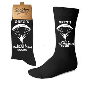 May include: Black socks featuring the text "GREG'S LUCKY PARAGLIDING SOCKS" in white, with a white paraglider graphic. The socks are presented against a white backdrop.