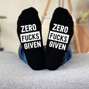 Może przedstawiać: Czarne skarpetki z białym napisem "ZERO FUCKS GIVEN". Skarpetki są noszone z niebieskimi dżinsami, spoczywając na szarej powierzchni. Humorystyczny przedmiot.