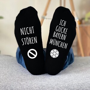 May include: Black socks with white text and graphics. One sock reads "NICHT ST&Ouml;REN" with a no-entry symbol, the other reads "ICH GUCKE BAYERN M&Uuml;NCHEN" with a soccer ball graphic. The socks are worn by someone.
