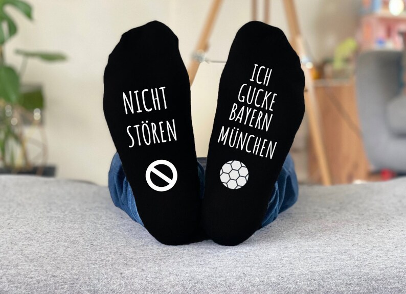 May include: Black socks with white text and graphics. One sock reads "NICHT ST&Ouml;REN" with a no-entry symbol, the other reads "ICH GUCKE BAYERN M&Uuml;NCHEN" with a soccer ball graphic. The socks are worn by someone.