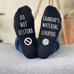 May include: Black socks with white text and graphics. One sock reads "DO NOT DISTURB" with a no-entry symbol. The other sock says "GRANDAD'S WATCHING LIVERPOOL" with a soccer ball graphic.