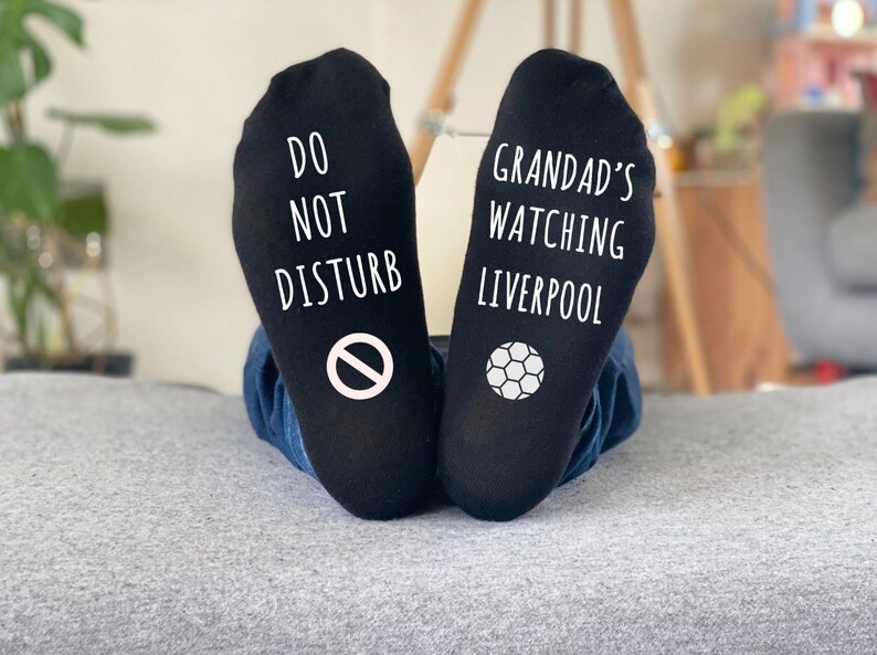May include: Black socks with white text and graphics. One sock reads "DO NOT DISTURB" with a no-entry symbol. The other sock says "GRANDAD'S WATCHING LIVERPOOL" with a soccer ball graphic.