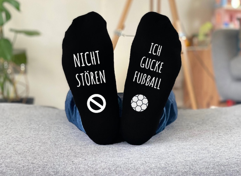 May include: Black socks with white text and graphics. One sock reads "NICHT ST&Ouml;REN" with a no-entry sign, the other reads "ICH GUCKE FUBBALL" with a soccer ball graphic. The socks are worn by someone wearing blue jeans.