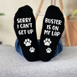 May include: Black socks with white text and paw prints. One sock says "SORRY I CAN'T GET UP" and the other says "BUSTER IS ON MY LAP". The socks are worn by someone in blue jeans.