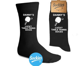 Könnte beinhalten: Schwarze Socken mit dem Text "DANNY'S LUCKY TABLE TENNIS SOCKS" und einer Grafik einer Tischtennisschlägers und eines Balls. Die Socken werden mit einem Pappanhänger mit der Aufschrift "Sockies" präsentiert. Ein blau-weißes "Sockies"-Logo ist ebenfalls sichtbar.