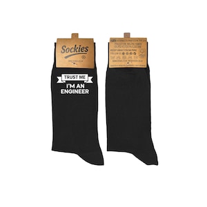 Puede incluir: Dos calcetines negros con el texto blanco "TRUST ME I'M AN ENGINEER". Cada calcetín tiene una etiqueta marrón con la marca "Sockies" e información sobre la composición y el cuidado de los calcetines.