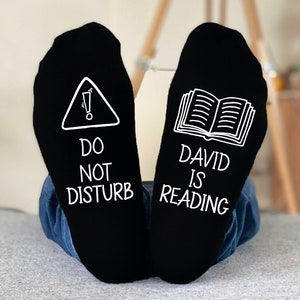 May include: Black socks with white text and graphics. One sock features a warning symbol and the words "DO NOT DISTURB." The other sock has an open book graphic and the words "DAVID IS READING."