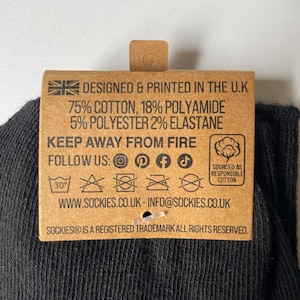 May include: A close-up of a brown tag attached to a black sock. The tag has text that reads "DESIGNED & PRINTED IN THE U.K" and details the sock's material composition. It also includes care instructions and social media icons.