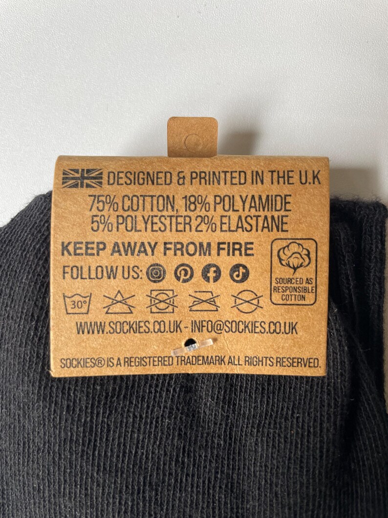 May include: A close-up of a brown tag attached to a black sock. The tag has text that reads "DESIGNED & PRINTED IN THE U.K" and details the sock's material composition. It also includes care instructions and social media icons.
