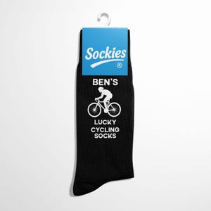 Pode incluir: Meias de ciclismo pretas com o texto "BEN'S LUCKY CYCLING SOCKS" e um gráfico branco de um ciclista. As meias têm uma etiqueta azul com a marca "Sockies".