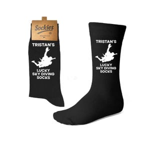 Puede incluir: Calcetines negros con el texto "TRISTAN'S LUCKY SKY DIVING SOCKS" en blanco. Los calcetines presentan un gráfico blanco de un paracaidista. Los calcetines se muestran sobre un fondo blanco.