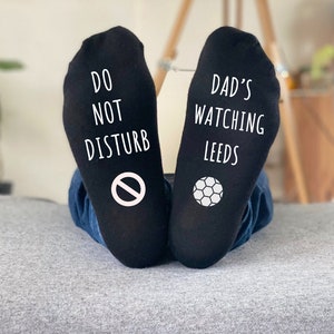 May include: Black socks with white text and graphics. One sock reads "DO NOT DISTURB" with a no-entry symbol. The other sock says "DAD'S WATCHING LEEDS" with a soccer ball graphic. The socks are worn by someone wearing blue jeans.