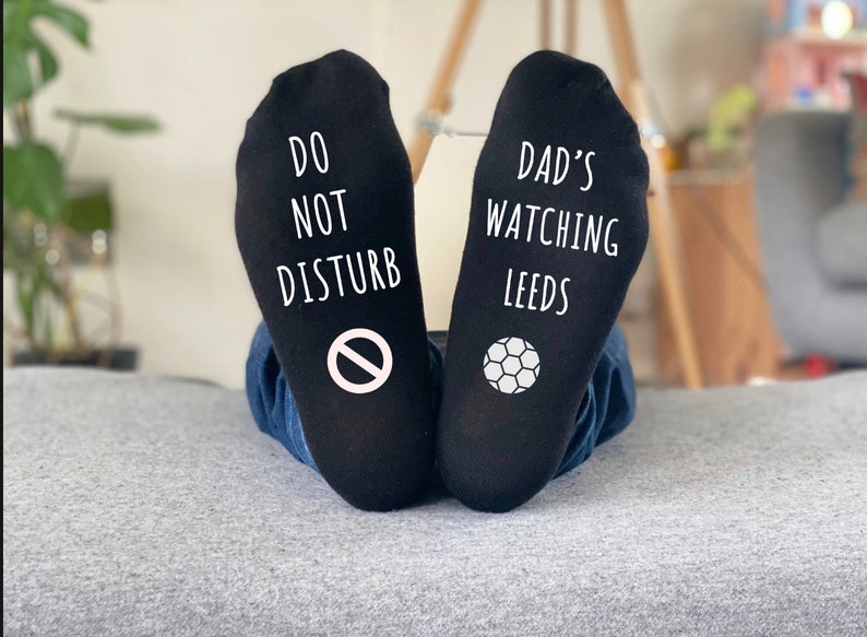May include: Black socks with white text and graphics. One sock reads "DO NOT DISTURB" with a no-entry symbol. The other sock says "DAD'S WATCHING LEEDS" with a soccer ball graphic. The socks are worn by someone wearing blue jeans.